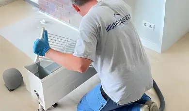 A cleaner from housecleaners.live kneels on the floor, deep cleaning the internal components of a radiator after removing the top grill.