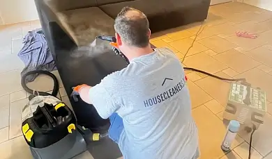 Professional cleaner from housecleaners.live steam cleaning the upholstery of a black sofa.