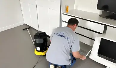 A cleaner from housecleaners.live kneels to clean the bottom shelf of a white wardrobe, with a large industrial vacuum standing next to him